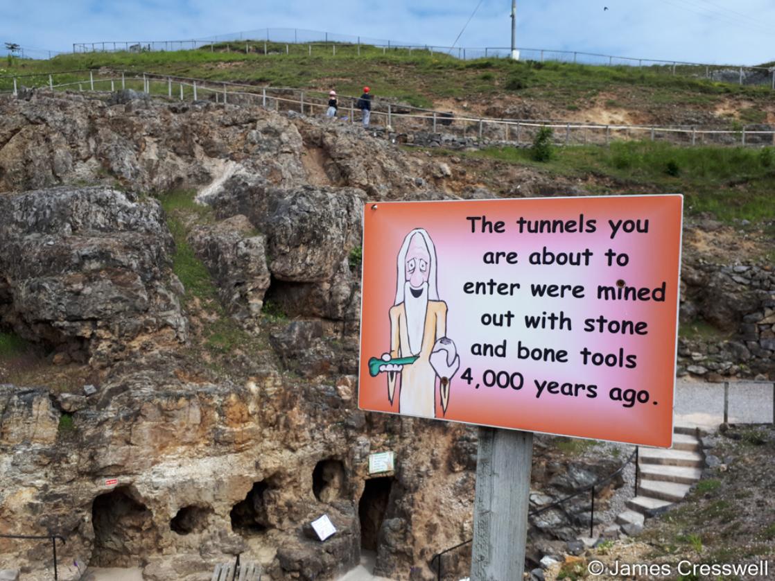 A photograph of a sign in front of a cliff face with tunnel openings, it is the Great Orme Bronze Age Copper Mine, taken on the GeoWorld Travel England and Wales geology trip, tour and holiday