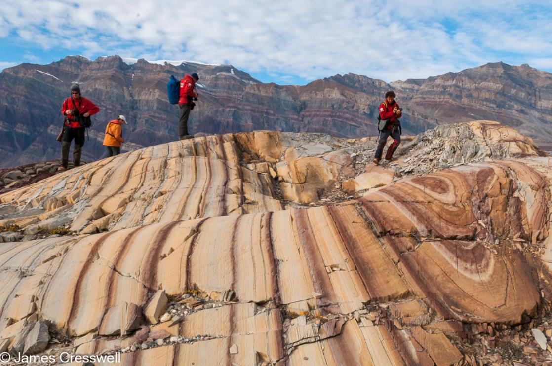 A photograph of people are standing on folded brown and white dolomite layers, in Segelsällskapets Fjord, northeast Greenland, with Berzelius Bjerg Mountain in the background. Taken on a PolarWorld Travel polar expedition cruise.
