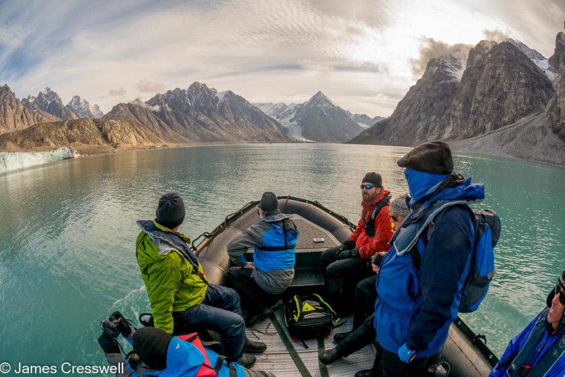 A photograph of a zodiac full of people passing down a fjord flanked with glaciers and pointed mountains, taken in Alpefjord in northeast Greenland on a PolarWorld Travel polar expedition cruise