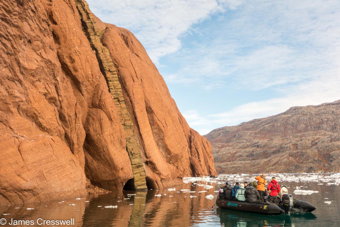 A photograph of people in a zodiac viewing a basaltic dyke cutting through bright red Permian aged sediments at Røde Ø, Scoresby Sund, east Greenland, take on a PolarWorld Travel polar expedition cruise