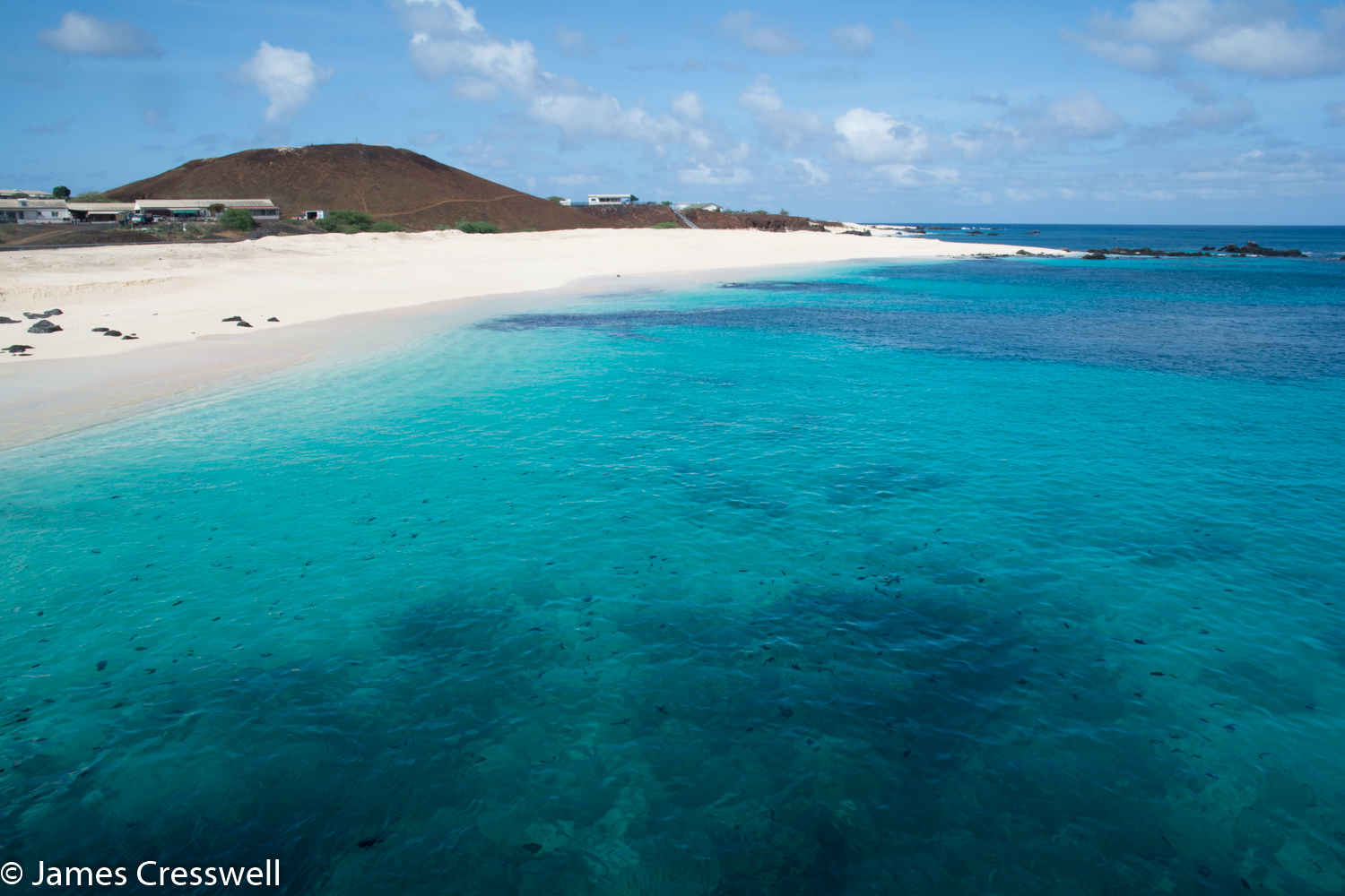 A photgraph of blue sea in the foreground, a white beach and red volcanic cone, Ascension Island, taken on a PolarWorld Travel placed cruise