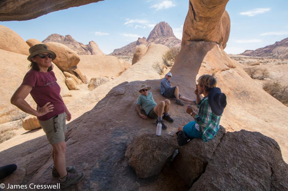A photograph of four people under a granite arch with Spitzkoppe mountain in the background, taken on a GeoWorld Travel Namibia geology trip, tour and holiday