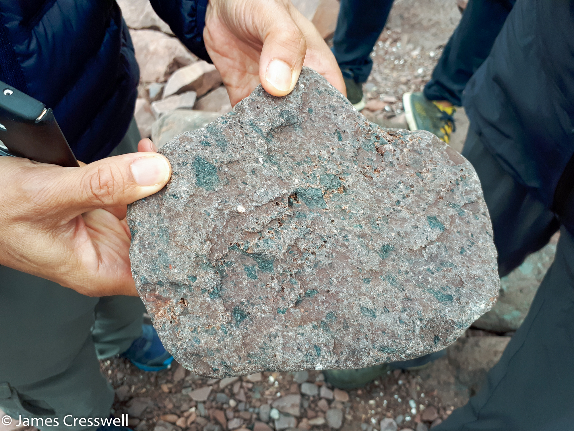 A photograph of a hand holding a reddish rock containing green glassy fragments, a one billion year old tektite at Stoer, taken on a GeoWorld Travel Scotland geology trip, tour and holiday