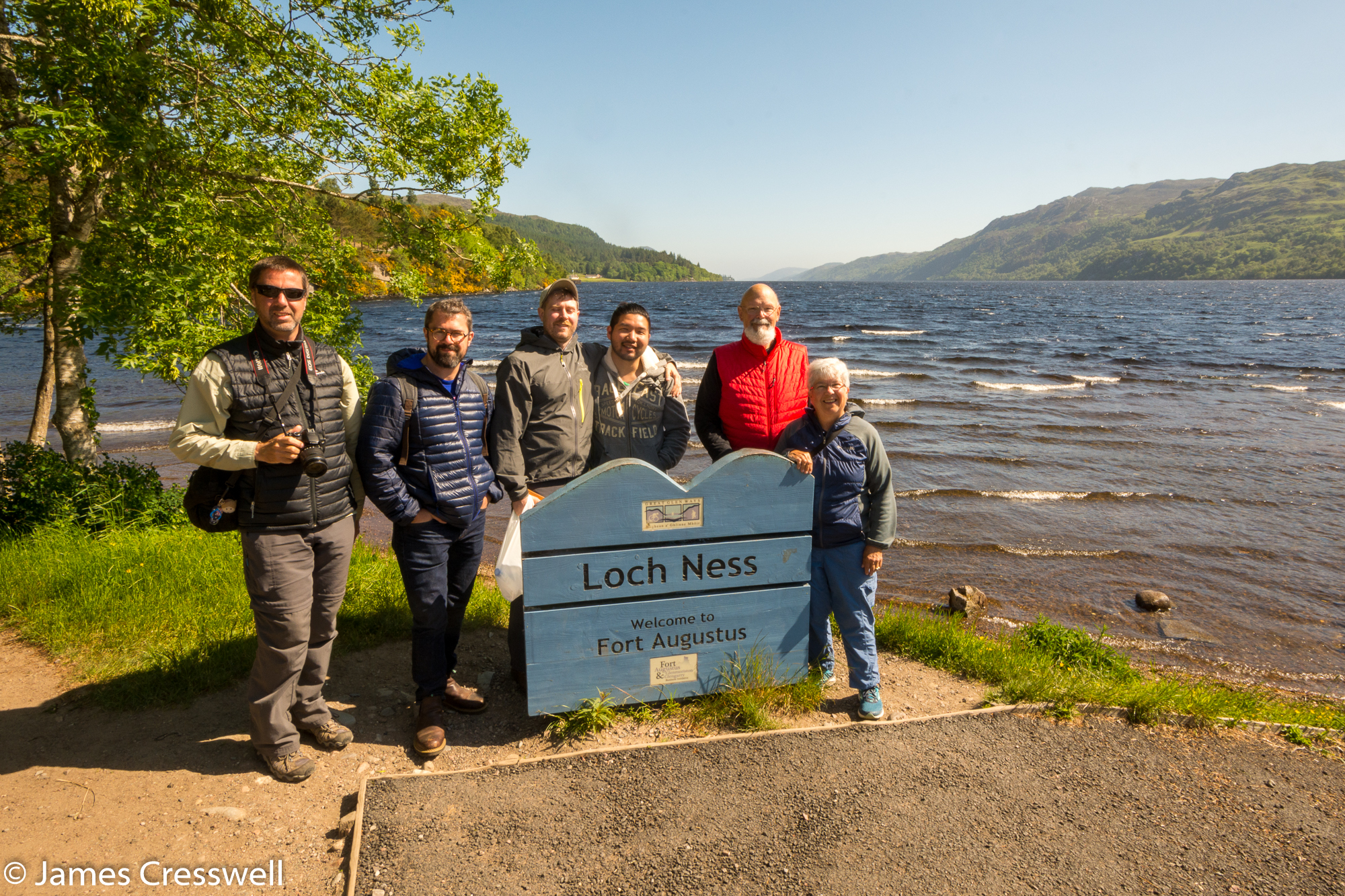 A photograph of six people standing next to the Loch Ness sign, with the loch in the background, taken on a GeoWorld Travel Scotland geology trip, tour and holiday
